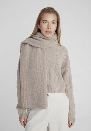Pull beige en maille avec texture torsadée et côtelée, assorti d'une écharpe de la même couleur. Tissu doux, coupe décontractée. Le mannequin porte un pantalon clair.