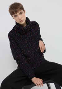 Maglione nero in maglia con colletto alto, caratterizzato da macchie colorate. Vestibilità ampia, tessuto testurizzato, indossato con pantaloni neri, seduto su una sedia.