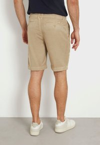 Pantalones cortos de algodón beige con puños enrollados, dos bolsillos traseros con botones y un acabado texturizado, combinados con zapatillas blancas sobre un suelo gris.