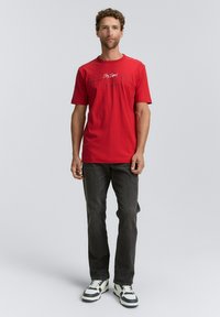 Rotes Baumwoll-T-Shirt mit kurzen Ärmeln und Rundhalsausschnitt, das vorne mit schwarzem Drucktext versehen ist. Kombiniert mit dunklen Jeans und Sneakern.