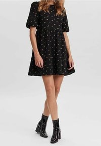 Schwarzes Kleid mit Puffärmeln, floralem Stickmuster, strukturiertem Stoff und ausgestelltem Rock. Kombiniert mit glänzenden schwarzen Stiefeletten.