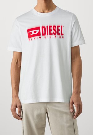 Mężczyzna ubrany w białą koszulkę Diesel z czerwonym logo i beżowe spodnie, stoi z rękami swobodnie opuszczonymi wzdłuż ciała.