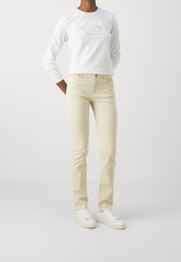 Vit långärmad sweatshirt med upphöjt logotyp, krämfärgade tajta jeans och vita sneakers på en modell mot en neutral bakgrund.