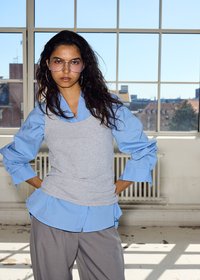 Femme aux longs cheveux foncés portant de grandes lunettes teintées, une chemise bleue, un gilet gris et un pantalon gris, debout à l'intérieur près d'une grande fenêtre avec vue sur la ville.