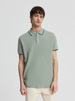 Młody mężczyzna ubrany w jasnooliwkową koszulkę polo z czarno-białym wykończeniem na kołnierzyku i rękawach, w połączeniu z jasnobeżowymi spodniami, stoi na jednolitym tle.