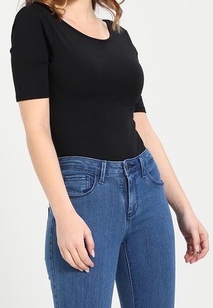 Kobieta ubrana w dopasowaną czarną bluzkę z okrągłym dekoltem i półdługimi rękawami oraz wysokim stanem niebieskie dżinsy, stoi z ramionami swobodnie opuszczonymi wzdłuż ciała.