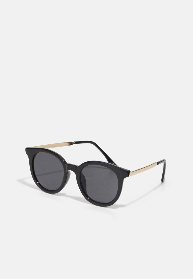 Óculos de sol pretos com armação redonda, lentes escuras e hastes finas com detalhes em dourado. A armação em plástico tem um acabamento suave e um design moderno.