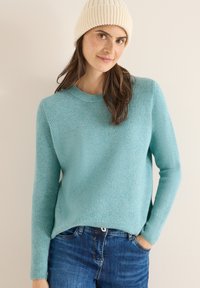 Hellblauer, gerippter Strickpullover mit langen Ärmeln und rundem Halsausschnitt, kombiniert mit dunkelblauen Jeans und einer crèmefarbenen Mütze.