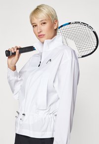Cilvēks ar īsiem blondiem matiem baltā žaketē tur tenisa raketi pār plecu uz vienkārša fona.