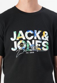 Black cotton t-shirt with "JACK & JONES Essentials" printed in colorful, textured letters. Crew neckline and short sleeves.