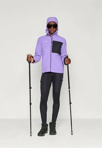 Svetlo fialová mikina na zips s čiernou náprsné kapsou, čierne priliehavé legíny, čierne turistické topánky a trekingové palice, nosené modelom.