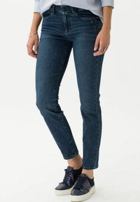 Jeans slim fit bleu foncé en denim, dotés d'un design classique à cinq poches et de détails de couture subtils, associés à des baskets noires et blanches.
