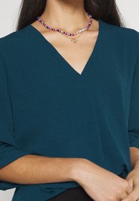 Top teal a V con maniche corte e una vestibilità ampia. Collana a più strati con perline colorate e un piccolo pendente a forma di cuore oro.