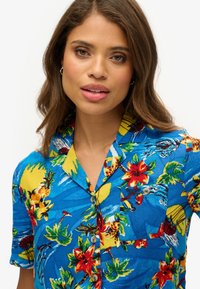 Camisa de manga corta de color azul con un patrón de flores tropicales en rojo y amarillo, con botones en el frente y un bolsillo en el pecho.
