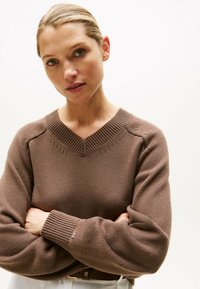 Brązowy sweter robiony na drutach z dekoltem w serek oraz ribbowymi mankietami. Ma teksturowany wzór i subtelne detale przeszycia wokół dekoltu.