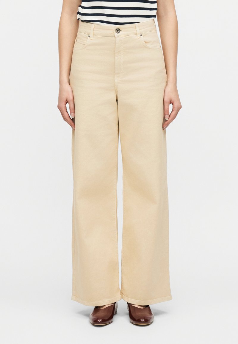 Γυναίκα που φοράει μπεζ παντελόνι  με φαρδιά πόδια, καφέ παπούτσια με τακούνι και ασπρόμαυρη ριγέ μπλούζα, στέκεται μπροστά σε απλό φόντο.