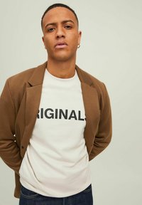 Brown blazer over a white shirt with "ORIGINALS" in bold black letters. Dark denim jeans visible at the waist. Neutral background.