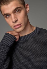 Dunkelgrauer strukturierter Pullover mit geripptem Rundhalsausschnitt. Das Material weist ein gestricktes Design und markante Nahtdetails an den Ärmeln auf.