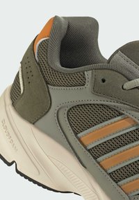Maslinasto zeleni sportski tenisice s mrežastim gornjim dijelom, odsušenim detaljima, narančastim prugama i bež Cloudfoam srednjim potplatom. Prikazani teksturirani materijali.