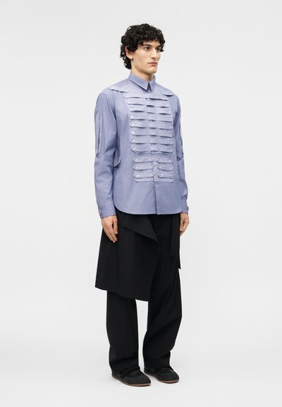 Jeune personne aux cheveux bouclés portant une chemise bleu clair à boutons avec des rabats superposés à l'avant et un pantalon noir large et ample.