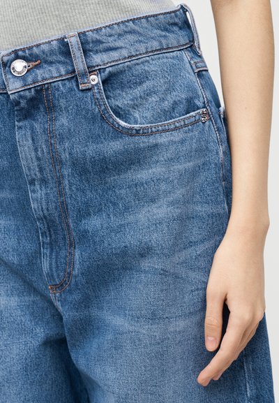 Κοντινό πλάνο μπλε τζιν με μεταλλικό κουμπί, μπροστινή τσέπη και ένα χέρι που ακουμπάει δίπλα στο ισχίο, φορώντας γκρι ριμπ μπλούζα.