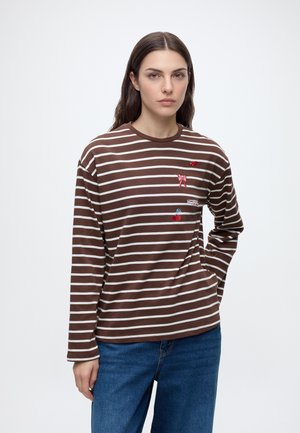 Vrouw met lang donker haar die een bruin-wit gestreept shirt met lange mouwen met kleine geborduurde motieven en een blauwe spijkerbroek draagt, staand tegen een witte achtergrond.