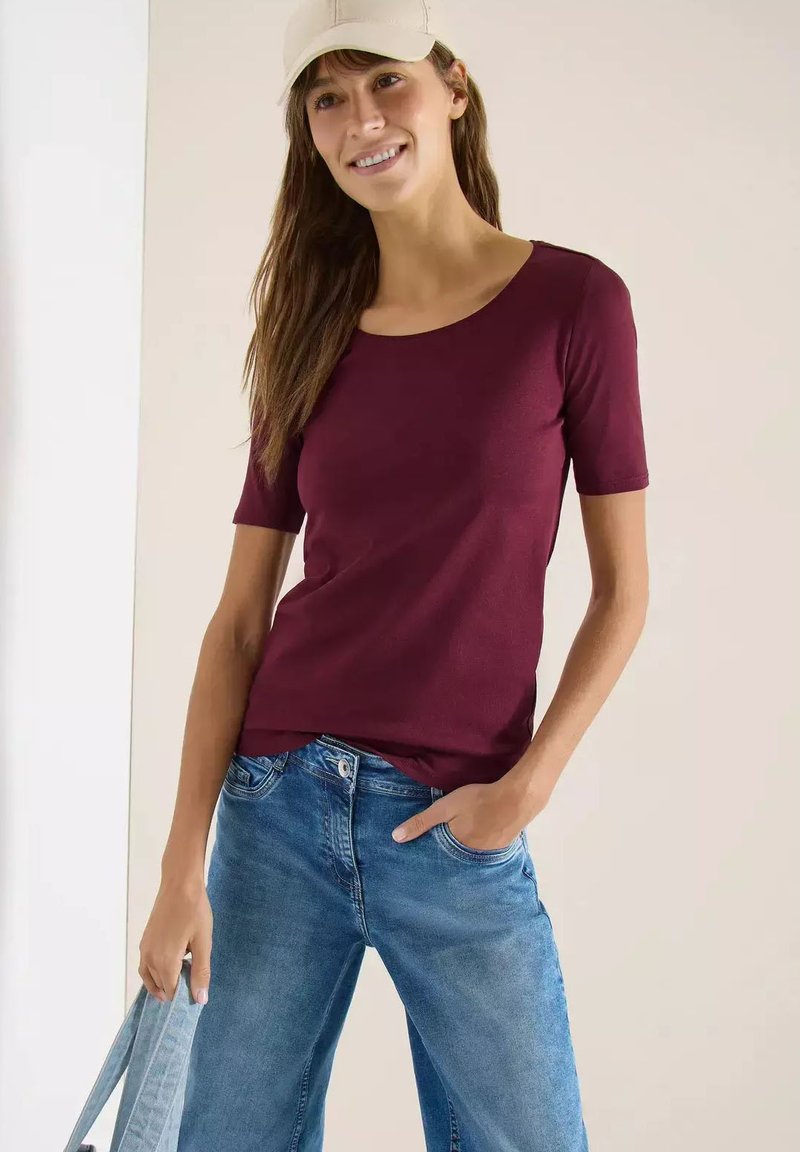 Femme portant une casquette beige, un haut manches courtes bordeaux et un jean bleu, tenant un sac bleu clair avec une main dans la poche.