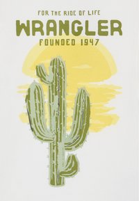 Hvit t-skjorte med et grønt kaktusmotiv, gult solbakgrunn og teksten "WRANGLER FOUNDED 1947" i fet grønn typografi.