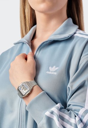 Sidabrinis skaitmeninis laikrodis su stačiakampiu ciferblatu, metaliniu dirželiu; mėlyna sportinė striukė su baltomis juostelėmis ir vyšninės siuvinėjimu ant krūtinės.