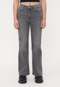 Jean évasé gris à taille haute, en denim. Comprend des poches avant, des passants de ceinture et une fermeture à bouton. Porté avec des chaussures massives noires.