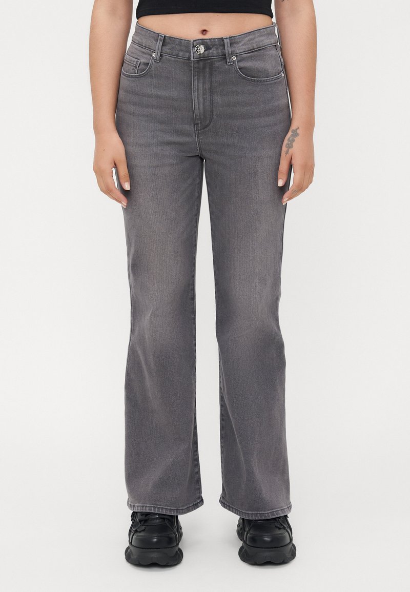 Jean évasé gris à taille haute, en denim. Comprend des poches avant, des passants de ceinture et une fermeture à bouton. Porté avec des chaussures massives noires.