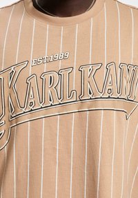 Dzeltenā svītrainā T-kreklā ir izteiksmīgi melna "KARL KANI" apdruka un „EST.1989” detaļa. Tas ir izgatavots no mīksta, viegla auduma ar apaļu kakla izgriezumu.