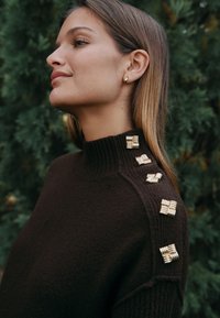 Mørkebrun strikket sweater med høj krave. Har gyldne metaldetaljer på skuldrene, som tilføjer tekstur og detaljer til designet.