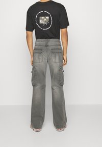 Graue Cargohosen mit weitem Bein, mehreren Taschen und Nahtdetails. Kombiniert mit einem schwarzen T-Shirt, das auf der Rückseite ein grafisches Design zeigt.