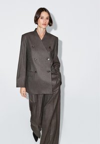 Šedý dvouřadý blaser s velkými knoflíky, dvěma předními kapsami a mírně oversize střihem, v kombinaci s odpovídajícími širokými kalhotami.
