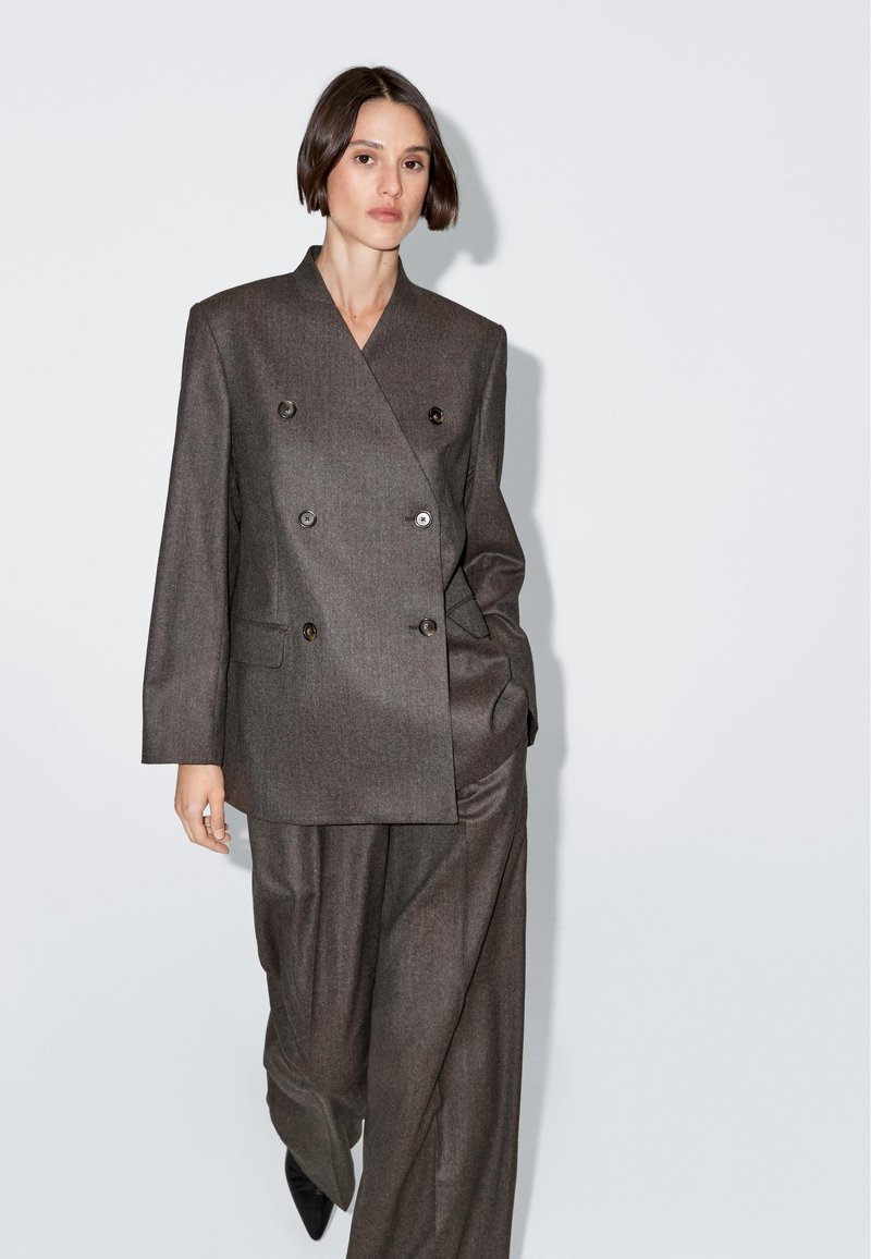 Šedý dvouřadý blaser s velkými knoflíky, dvěma předními kapsami a mírně oversize střihem, v kombinaci s odpovídajícími širokými kalhotami.