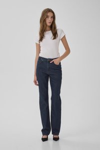 Mladá žena s dlhými vlasmi oblečená v priliehavom bielom tričku s krátkym rukávom, tmavomodrých džínsoch a čiernych špicatých lodičkách, stojaca na pozadí bez vzoru.