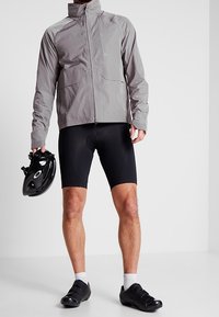 Veste de cyclisme légère grise avec un col montant, deux poches avant et un short de cyclisme noir. Casque noir tenu dans la main gauche, chaussures noires élégantes.