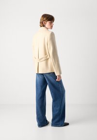 Bežový strihový blejzer s rozparkom a mašľou, nosený cez voľné modré denimové džínsy, na jednofarebnom bielom pozadí.