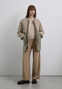 Chaqueta beige acolchada con textura suave, paneles verdes en la parte inferior y botones de presión. Combinada con pantalones anchos color marrón claro y botas de plataforma marrones.