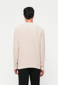 Homme portant un pull à manches longues texturé beige et un pantalon noir, se tenant face à un mur blanc, vu de derrière.