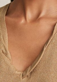 Złoty sweter z dekoltem w serek, charakteryzujący się metalicznym połyskiem, miękką fakturą i luźnym fasonem, z widocznymi szwami i głębokim dekoltem.