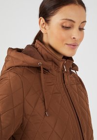 Giacca marrone trapuntata con cappuccio, caratterizzata da un colletto alto, dettagli concordabili e una texture liscia. Chiusura a zip sul davanti.