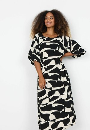 Ženska z kodrastimi lasmi, ki se smeji, oblečena v črno-belo vzorčasto midi obleko s puhastimi rokavi, stoji pred belim ozadjem.