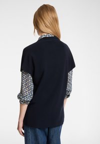 Frau mit langen blonden Haaren, die einen marineblauen kurzärmligen Pullover über einer blauen gemusterten Bluse und blauen Jeans trägt, von hinten gesehen.