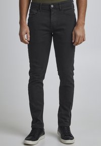 Jeans de mezclilla negra con corte ajustado, que cuentan con cinco bolsillos, cierre de botón y una ligera textura. Combinados con zapatillas negras.