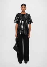 Black sequined oversized top with short sleeves, paired with wide-leg black pants. The model carries a black satchel bag.