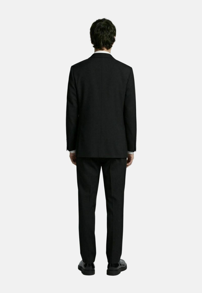 Hombre de espaldas, vestido con un traje negro con pantalones a juego y zapatos de vestir negros sobre un fondo claro y liso.