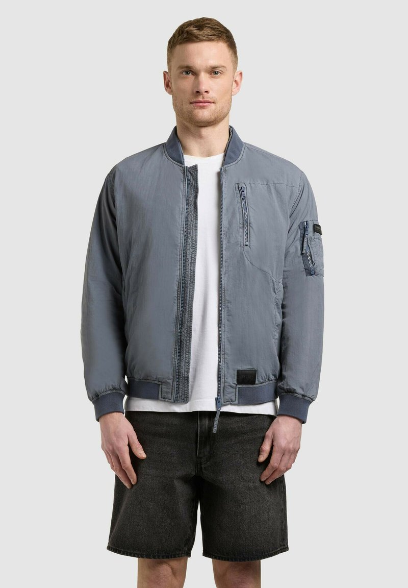 Homme portant une veste bomber grise sur une chemise blanche et un short en jean noir, debout devant un fond clair uni.