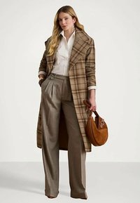 Γυναίκα που φορά καφέ καρό παλτό, λευκό πουκάμισο, παντελόνι με φαρδιά πόδια και ψηλή μέση, κρατώντας μία καφέ σουέτ τσάντα χεριού μπροστά σε απλή φόντο.